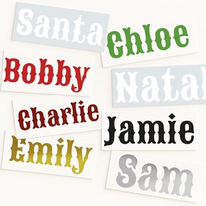 May include: A collection of name decals in various colors and fonts. Names include "Santa", "Chloe", "Bobby", "Natalie", "Charlie", "Jamie", "Emily", and "Sam". The decals are on rectangular backgrounds.