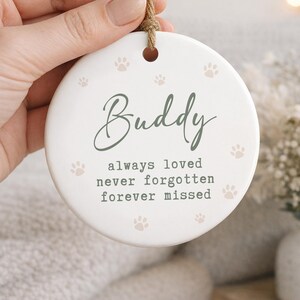 May include: A white ceramic ornament with the name "Buddy" in green script, surrounded by paw prints. The text "always loved, never forgotten, forever missed" is below the name. A jute string is attached for hanging.