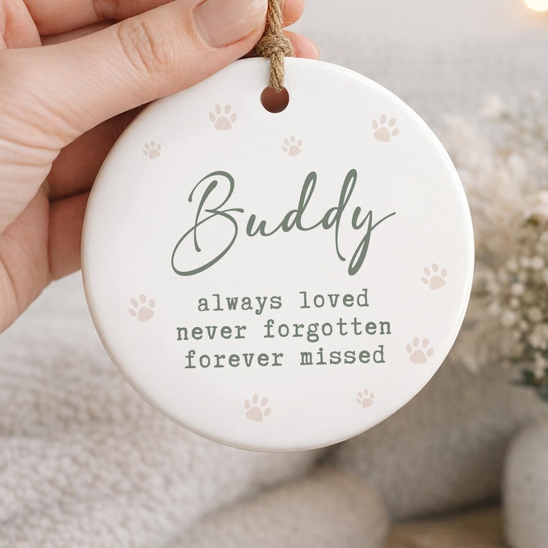 May include: A white ceramic ornament with the name "Buddy" in green script, surrounded by paw prints. The text "always loved, never forgotten, forever missed" is below the name. A jute string is attached for hanging.