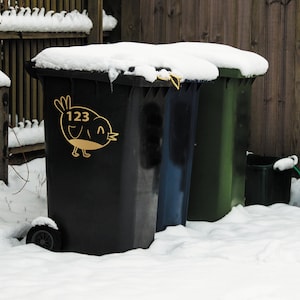 K&ouml;nnte beinhalten: Drei schwarze, blaue und gr&uuml;ne Recyclingbeh&auml;lter, die mit Schnee bedeckt sind, mit der Nummer 123 und einem Vogeldesign auf dem schwarzen Beh&auml;lter.