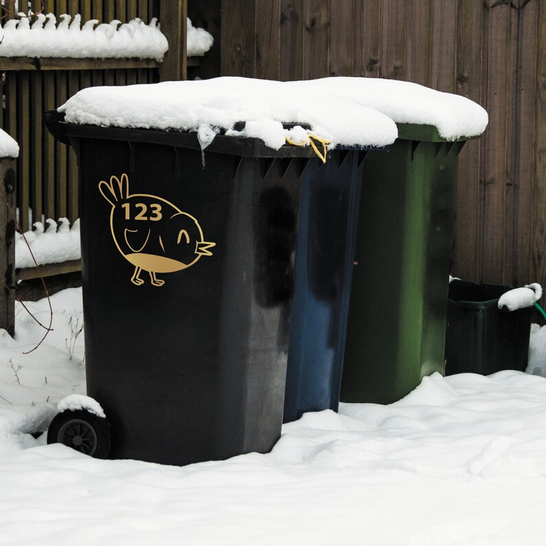 K&ouml;nnte beinhalten: Drei schwarze, blaue und gr&uuml;ne Recyclingbeh&auml;lter, die mit Schnee bedeckt sind, mit der Nummer 123 und einem Vogeldesign auf dem schwarzen Beh&auml;lter.
