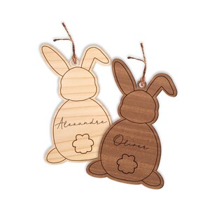 May include: Two wooden bunny-shaped ornaments with names "Alexandre" and "Oliver" engraved on them. The bunnies are brown and light brown, with a small flower design on their tails.