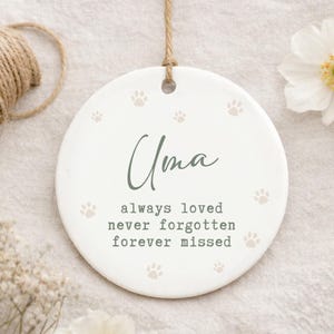 May include: A white ceramic ornament with the name "Uma" in green script, surrounded by paw prints. The text "always loved, never forgotten, forever missed" is below the name. A jute string is attached for hanging.