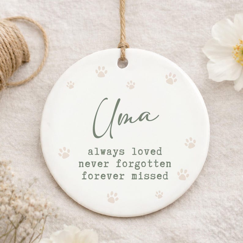 May include: A white ceramic ornament with the name "Uma" in green script, surrounded by paw prints. The text "always loved, never forgotten, forever missed" is below the name. A jute string is attached for hanging.