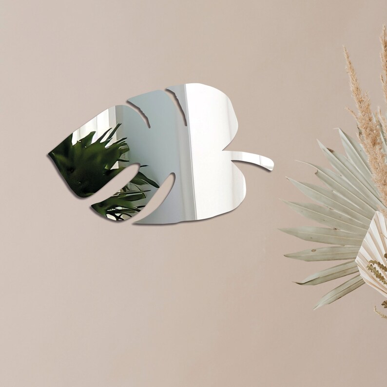 Pu&ograve; includere: Uno specchio argentato a forma di foglia di monstera. Lo specchio &egrave; montato su una parete color beige chiaro.