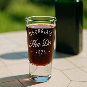 May include: A clear shot glass filled with a dark liquid, featuring the text "GEORGIA'S Hen Do - 2025" in a white, elegant font. A dark green bottle is slightly visible in the background. The glass sits on a light-colored surface.