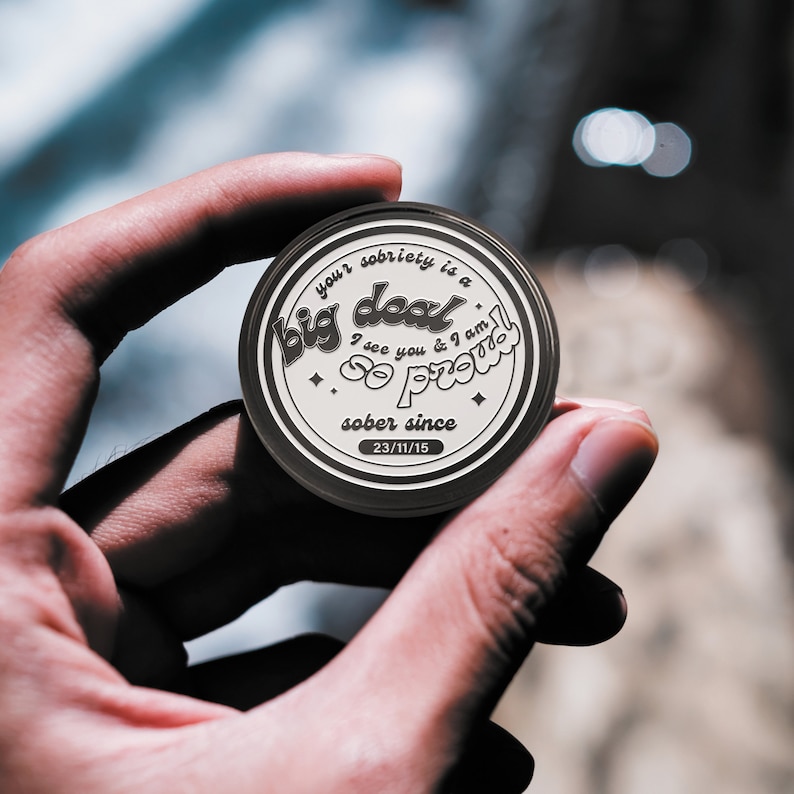 May include: A round, black and white sobriety token with the text "your sobriety is a big deal I see you & I am so proud sober since 23/11/15".