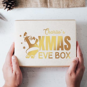 May include: A brown cardboard box with a gold foil design featuring a cartoon Santa Claus and the text "Charlie's KMAS EVE BOX".