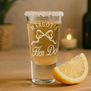 May include: A clear shot glass with a salted rim, filled with a golden liquid. The glass is etched with the words "Margot's Hen Do" and a bow design. A lemon wedge rests beside the glass.