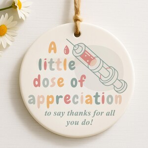 May include: A round, off-white ceramic ornament with a syringe illustration and the text "A little dose of appreciation to say thanks for all you do!" The syringe is red and gray, and the text is in various pastel colors.