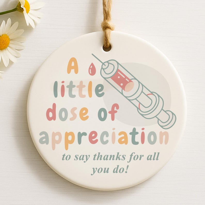 May include: A round, off-white ceramic ornament with a syringe illustration and the text "A little dose of appreciation to say thanks for all you do!" The syringe is red and gray, and the text is in various pastel colors.
