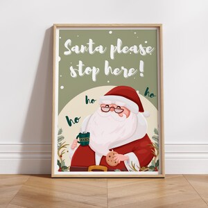 May include: A printable Christmas sign featuring a cartoon Santa Claus holding a cup of hot cocoa and a cookie. The sign says "Santa please stop here!" with "ho ho ho" written around Santa.