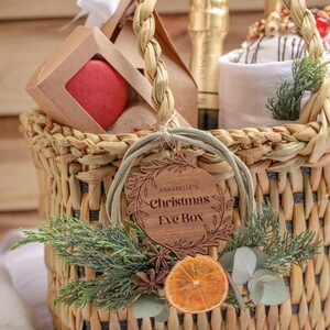 以下が含まれることがあります： クリスマスイブの祝賀会のためのギフトが詰まった籐のバスケット。バスケットは緑の葉、乾燥したオレンジのスライス、そして「Annabelle's Christmas Eve Box」と書かれた木製のオーナメントで飾られています。