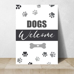 May include: A white sign with the words "DOGS Welcome" in black and gray. The sign features paw prints and a bone graphic with a heart. The sign is designed to welcome dogs.