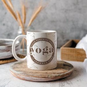 K&ouml;nnte beinhalten: Wei&szlig;e Keramik-Tasse mit braunem, kreisf&ouml;rmigem Design mit der Aufschrift "Yoga is frowned upon because punching people". Die Tasse hat einen Henkel und ist mit Yoga-Posen verziert. Die Tasse steht auf einem Holzuntersetzer.