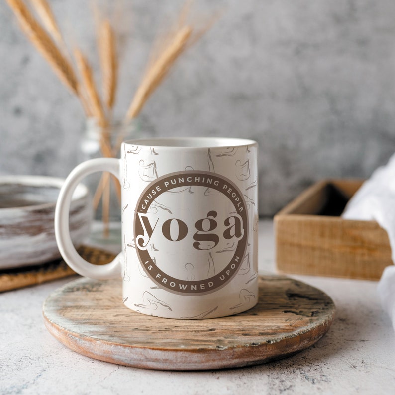 K&ouml;nnte beinhalten: Wei&szlig;e Keramik-Tasse mit braunem, kreisf&ouml;rmigem Design mit der Aufschrift "Yoga is frowned upon because punching people". Die Tasse hat einen Henkel und ist mit Yoga-Posen verziert. Die Tasse steht auf einem Holzuntersetzer.