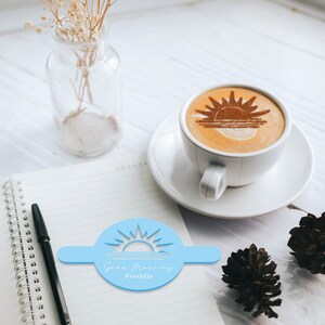 May include: A blue plastic stencil with a sunrise design and the text "Good Morning Freddie". The stencil is on a white notebook with a black pen. A cup of coffee with a cinnamon sunrise design is in the background.