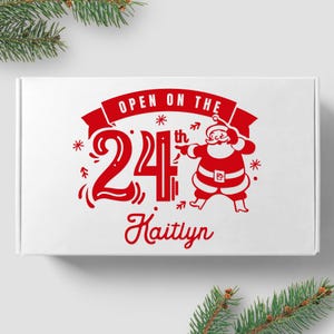 May include: White box with red text and a red illustration of Santa Claus. The text reads "Open on the 24th Kaitlyn".