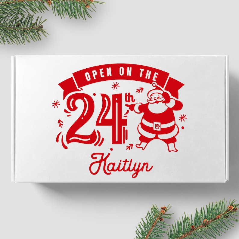 May include: White box with red text and a red illustration of Santa Claus. The text reads "Open on the 24th Kaitlyn".