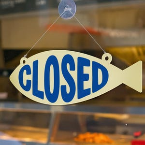 May include: A cream-colored, fish-shaped sign with the word "CLOSED" in bold, blue letters. The sign is suspended by a string from a suction cup attached to a glass surface. The background is blurred, suggesting a shop interior.