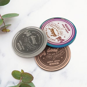 May include: Three round metal tokens with the text "Big Deal" and "So Proud" and the date "sober since" followed by a date. The tokens are silver, copper, and a rainbow color.