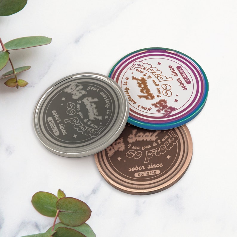 May include: Three round metal tokens with the text "Big Deal" and "So Proud" and the date "sober since" followed by a date. The tokens are silver, copper, and a rainbow color.