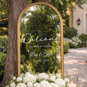 May include: A gold-framed mirror with an arched top, displaying the words "Welcome TO OUR WEDDING Ben & Kate 22/07/2026" in elegant white script. The mirror is surrounded by white flowers, with a blurred outdoor background.