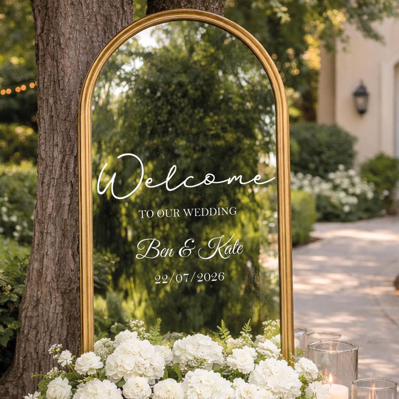 May include: A gold-framed mirror with an arched top, displaying the words "Welcome TO OUR WEDDING Ben & Kate 22/07/2026" in elegant white script. The mirror is surrounded by white flowers, with a blurred outdoor background.