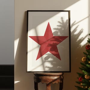 May include: Framed art print featuring a large, textured red star on a white background. The artwork is displayed in a black frame and rests on a dark wooden table. The image is lit by natural light, with shadows from a nearby tree.