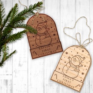 May include: Two wooden Christmas decorations shaped like snow globes with a snowman inside. The decorations are personalised with the text "Kaitlin's First Christmas" and "Emma's First Christmas".