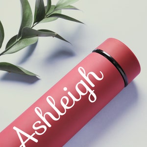 K&ouml;nnte beinhalten: Eine rosafarbene Wasserflasche aus Edelstahl mit einer wei&szlig;en Aufschrift "Ashleigh".