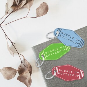 Puede incluir: Tres llaveros con el texto "Buckle Up Buttercup" en blanco sobre fondo azul, verde y rojo. Los llaveros tienen forma de llaves de motel.