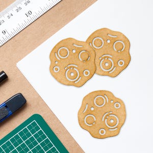 Puede incluir: Tres recortes de madera con forma de planetas con dise&ntilde;os circulares. Los recortes son de color marr&oacute;n claro y tienen una textura rugosa.