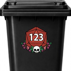 May include: Black trash can with a red, white, and green graphic. The graphic features a red polygon with the number 123, surrounded by flowers and a skull.