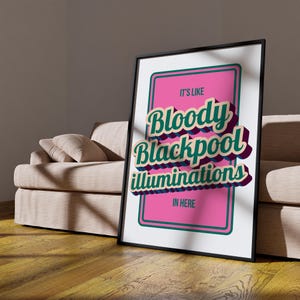 以下が含まれることがあります： 「It's Like Bloody Blackpool Illuminations In Here」のテキストが入ったフレーム付きポスター。テキストは3Dスタイルで、ティールと紫のアクセントが施され、ピンクの背景に配置されています。フレームは黒で、ポスターはベージュのソファに立てかけられています。