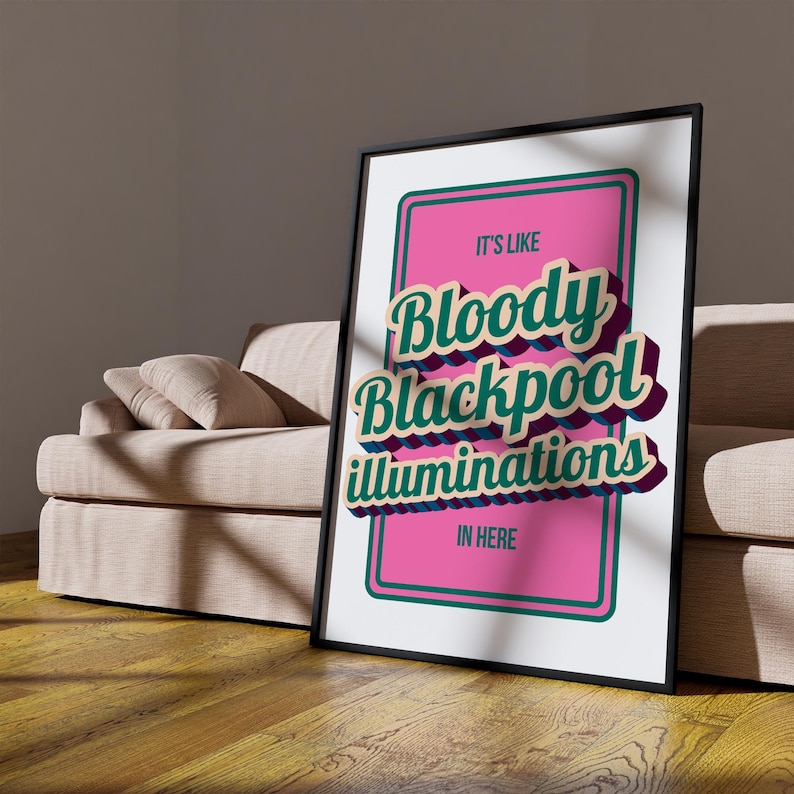 以下が含まれることがあります： 「It's Like Bloody Blackpool Illuminations In Here」のテキストが入ったフレーム付きポスター。テキストは3Dスタイルで、ティールと紫のアクセントが施され、ピンクの背景に配置されています。フレームは黒で、ポスターはベージュのソファに立てかけられています。