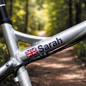 May include: Close-up of a silver bicycle frame with the name "Sarah" in dark blue lettering. A small Union Jack flag sticker is positioned next to the name. Black cables and a black handlebar are also visible.