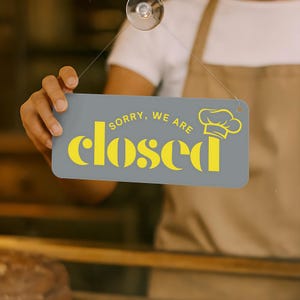 Peut inclure: Un panneau rectangulaire gris avec les mots "SORRY, WE ARE closed" en jaune, et une ic&ocirc;ne de toque de chef. Le panneau est suspendu sur une surface vitr&eacute;e.