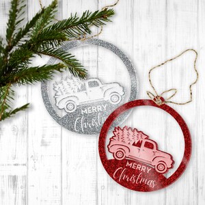 May include: Two round Christmas ornaments with a red truck carrying a Christmas tree. The ornaments are silver and red with glitter. The silver ornament says "Merry Christmas" and the red ornament says "Merry Christmas".