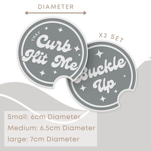 May include: Set of two gray round car coasters with white text. One coaster says "That Curb Hit Me" and the other says "Buckle Up".