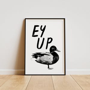 Op de afbeelding: Zwart-witte print van een eend met de tekst "Ey Up" in een zwart frame.