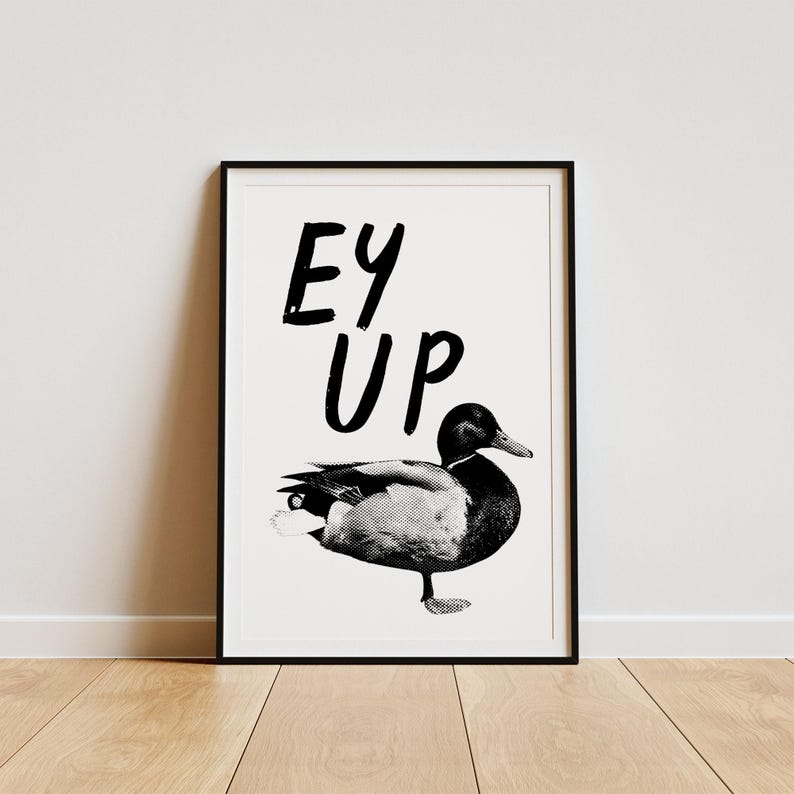 Op de afbeelding: Zwart-witte print van een eend met de tekst "Ey Up" in een zwart frame.