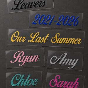 May include: A collection of clear acrylic name tags and labels with various text in different colors. The labels include the words "Leavers", "2021-2026", "Our Last Summer", and names like "Ryan", "Amy", "Chloe", and "Sarah".