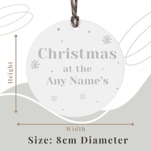 Peut inclure: Ornement de No&euml;l circulaire blanc avec le texte "Christmas at the Any Name's" en gris. L'ornement pr&eacute;sente des motifs de flocons de neige et de points, et une ficelle marron pour l'accrocher. Le diam&egrave;tre est de 8 cm.