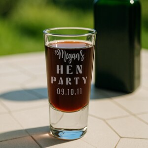 Op de afbeelding: Een helder shotglaasje gevuld met een donkere vloeistof. Op het glas staat in witte letters "Megans HEN PARTY 09.10.11". Op de achtergrond is een donkergroene fles te zien, wat duidt op een feest of viering.