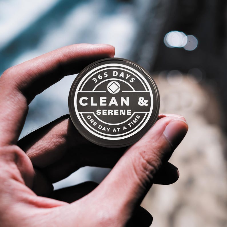 May include: A black round token with white text that reads "365 Days Clean & Serene One Day At A Time". The token has a diamond shape in the center.