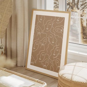 Op de afbeelding: Een ingelijst print met een minimalistisch ontwerp met meerdere abstracte gezichten in witte contouren op een bruine achtergrond.