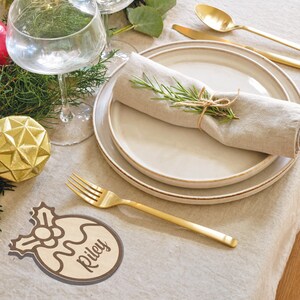 Pu&ograve; includere: Una mise en place natalizia con un segnaposto in legno su cui &egrave; scritto "Riley". La carta &egrave; a forma di ornamento natalizio con un ramo di agrifoglio e una bacca rossa. La mise en place comprende anche un tovagliolo bianco con un rametto di rosmarino, una forchetta dorata e un piatto bianco.