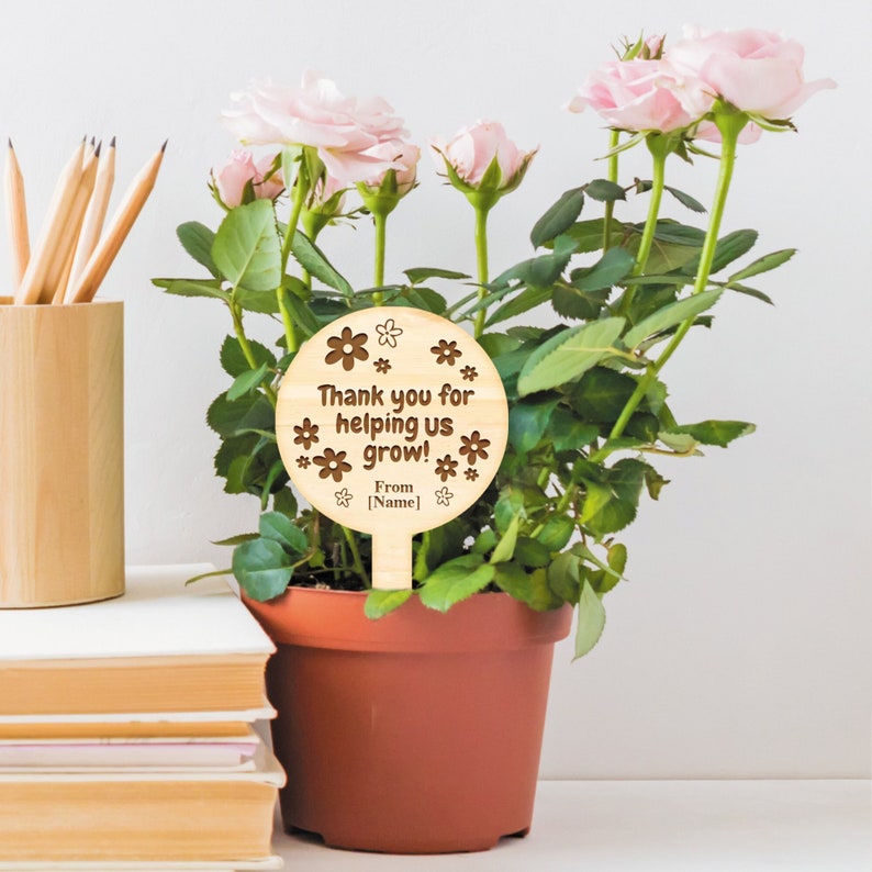May include: Wooden plant stake with a round sign that says "Thank you for helping us grow! From [Name]" with a flower design. The stake is in a pot with a pink rose bush.