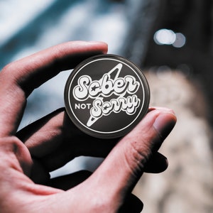 May include: A black round pin with a white lightning bolt and the text "Sober Not Sorry" in a retro font.